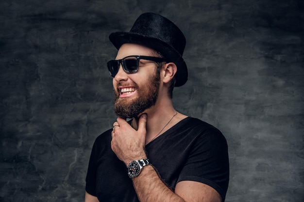 Free photo portrait of bearded male in a black t shirt, cylinder hat and sunglasses over grey background.