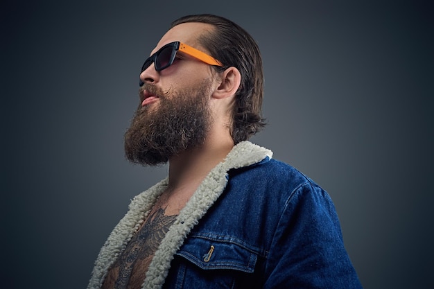 Free photo portrait of bearded male in sunglasses with tattoo on a chest wearing a denim jacket.