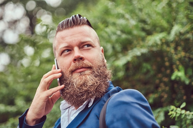 Free photo portrait of a bearded male with a haircut dressed in a shirt and jacket with a backpack, talking by phone in a park.