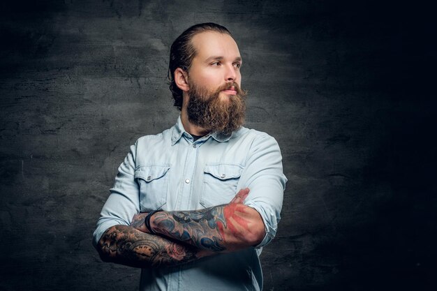 Portrait of bearded male with tattooed crossed arms on grey background.