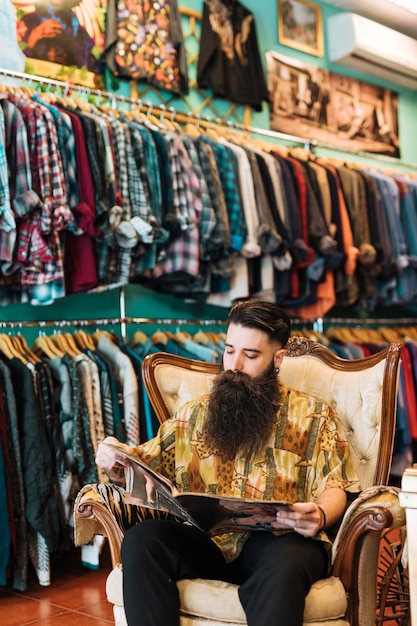 Free photo portrait of a bearded man sitting on antique arm chair looking at magazine in clothes shop