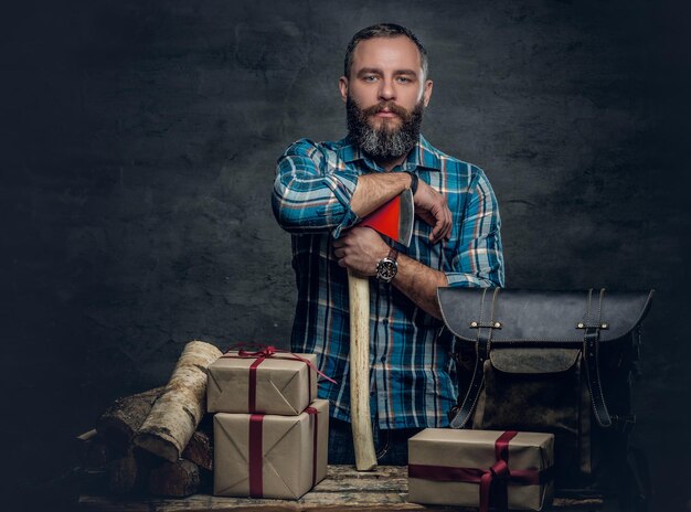 Portrait of bearded middle age male holds an axe and standing near a table with Christmas gift boxes and firewoods over grey background.