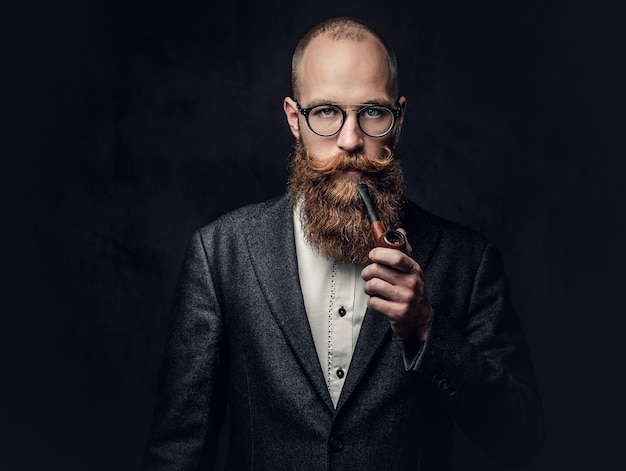 Portrait of bearded redhead English male smoking pipe over grey background.