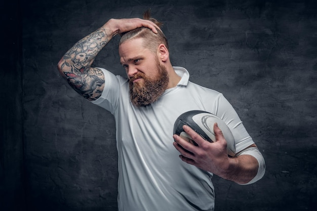 Portrait of bearded rugby player with tattoos on his arms holds a game ball.