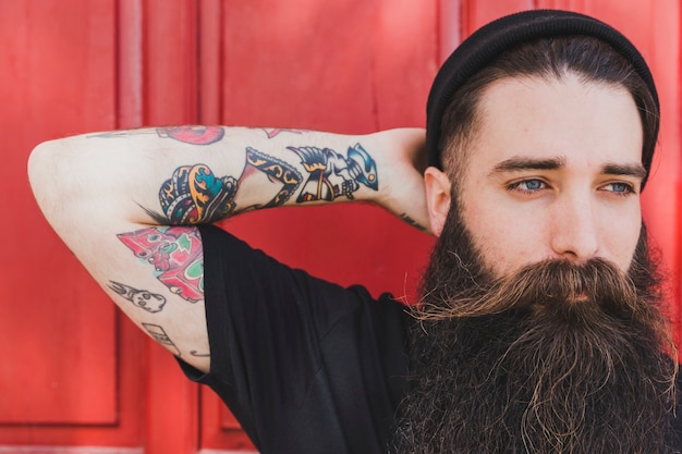 Portrait of a bearded young man with colorful tattoo on his hand against red backdrop