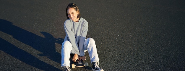 Free photo portrait of beautiful asian girl skating sitting on her skateboard and smiling cute teenager with