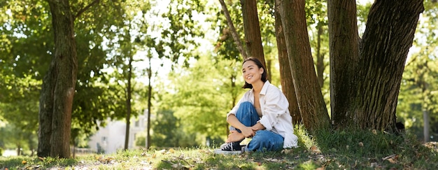 Free photo portrait of beautiful asian woman resting near tree relaxing in park smiling and looking happy