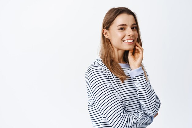 Portrait of beautiful blond girl touching face and smiling sincere, showing pure glowing skin without make up, standing over white