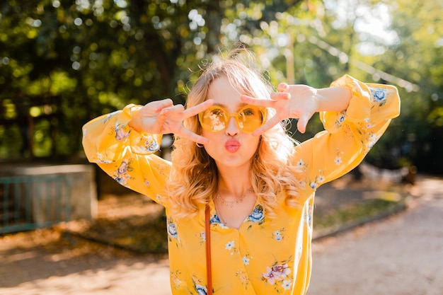 Portrait of beautiful blond stylish emotional woman in yellow blouse wearing sunglasses, funny crazy face expression