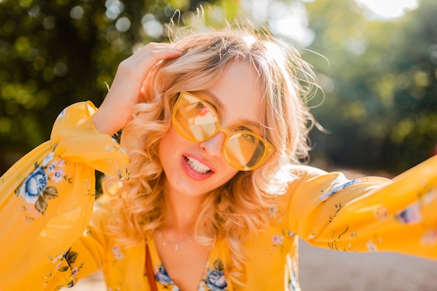 Portrait of beautiful blond stylish  smiling woman in yellow blouse wearing sunglasses making selfie photo