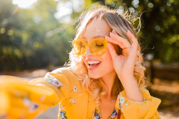 Free photo portrait of beautiful blond stylish smiling woman in yellow blouse wearing sunglasses making selfie photo