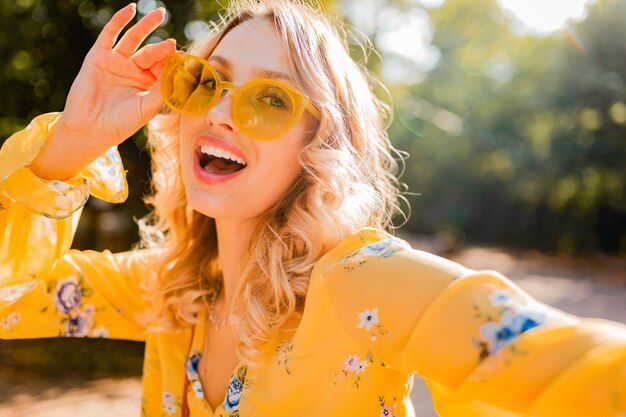 Portrait of beautiful blond stylish smiling woman in yellow blouse wearing sunglasses making selfie photo