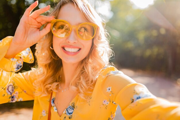 Portrait of beautiful blond stylish smiling woman in yellow blouse wearing sunglasses making selfie photo