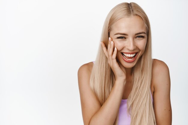 Portrait of beautiful blond woman smiling laughing and touching shiny soft facial skin standing over white background Copy space