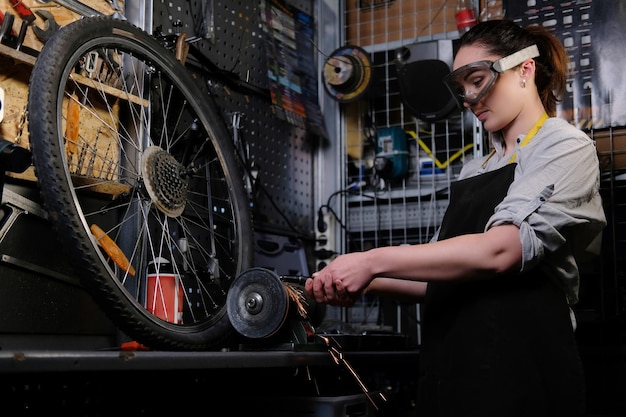 Portrait of a beautiful brunette female wearing working clothes, apron and goggles, working on a sharpening machine in a workshop.