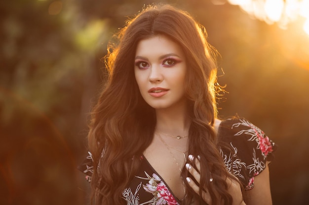 Free photo portrait of a beautiful brunette girl with wavy thick hair against a city sunset a girl in short jeans shorts and a black colorful blouse with stylish makeup and jewelry