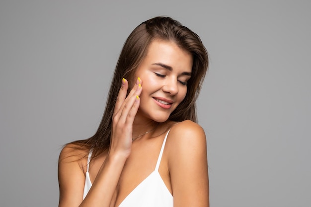 Portrait of beautiful brunette with bare shoulders touching her cheek, looking at camera and smiling on gray