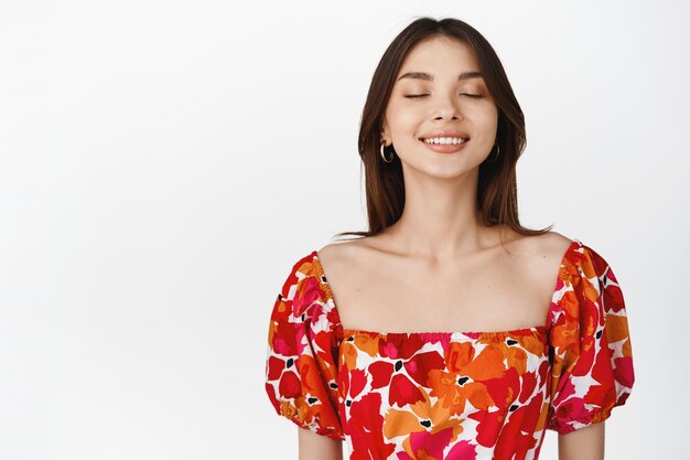 Portrait of beautiful brunette woman close eyes and dreaming smiling while imaging something standing in floral dress over white background