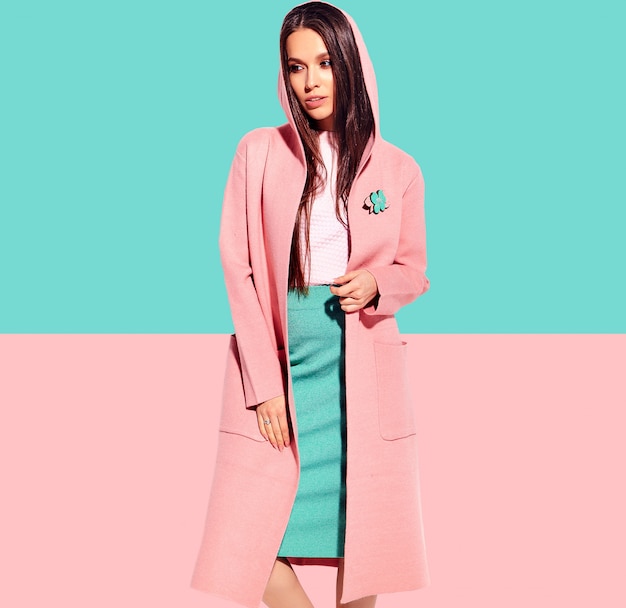 Portrait of beautiful caucasian smiling brunette woman model in bright overcoat and summer stylish skirt posing  on pink and blue background