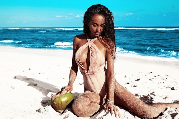 portrait of beautiful caucasian sunbathed woman model with dark long hair in beige swimsuit posing on summer beach with white sand on blue sky and ocean