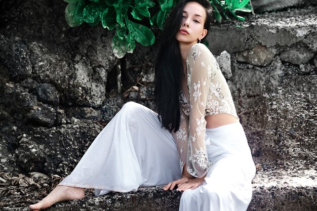 Free Photo portrait of beautiful caucasian woman model with dark long hair in wide-leg classic pants  posing near rocks and green tropical exotic leaves background
