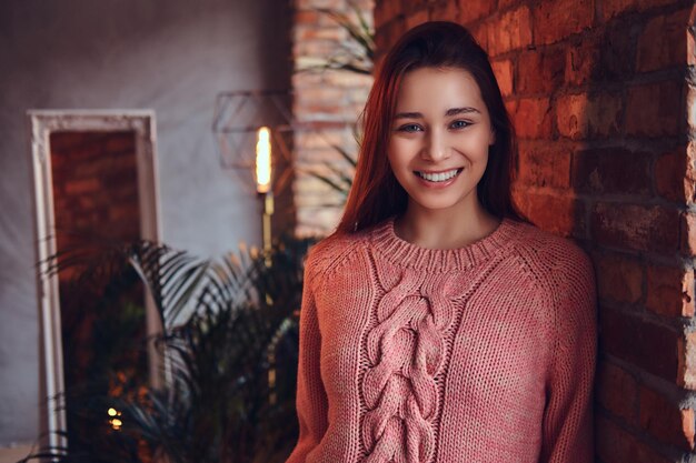 Portrait of a beautiful charming brunette dressed in stylish clothes smiling while leaning against a brick wall in a room with loft interior.