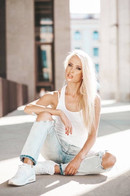 Free Photo portrait of beautiful cute blond girl in white t-shirt and jeans posing outdoors. cute girl sitting on asphalt on the street