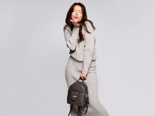 Portrait of beautiful cute brunette woman model in casual autumn gray sweater clothes with no makeup isolated on gray with handbag. GIving a kiss