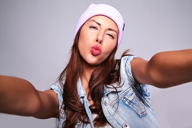 Portrait of beautiful cute brunette woman model in casual summer jeans clothes with no makeup in purple beanie making selfie photo on phone isolated on gray. Giving air kiss