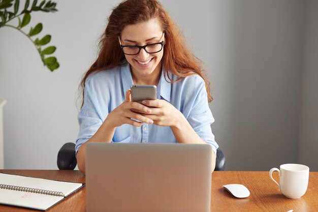 Portrait of beautiful female secretary wearing blue shirt and glasses, smiling while texting