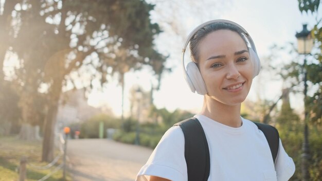 Portrait of beautiful girl in headphones with backpack smiling on camera on the way to morning training in city park