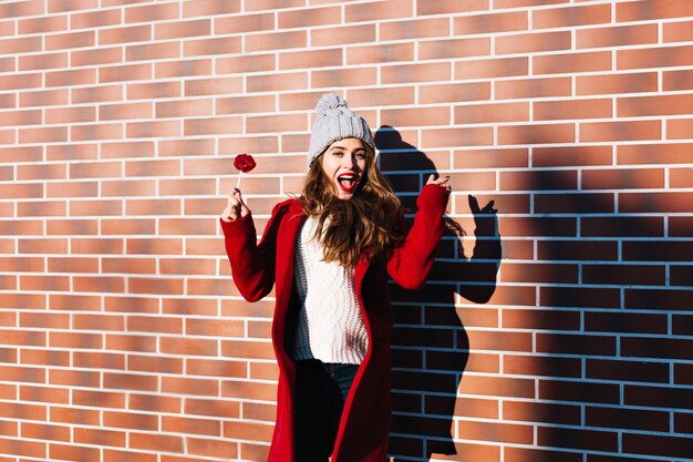 Free Photo portrait beautiful girl in red coat with lollipop lips on wall  outside. she wears knitted hat, looks satisfied .