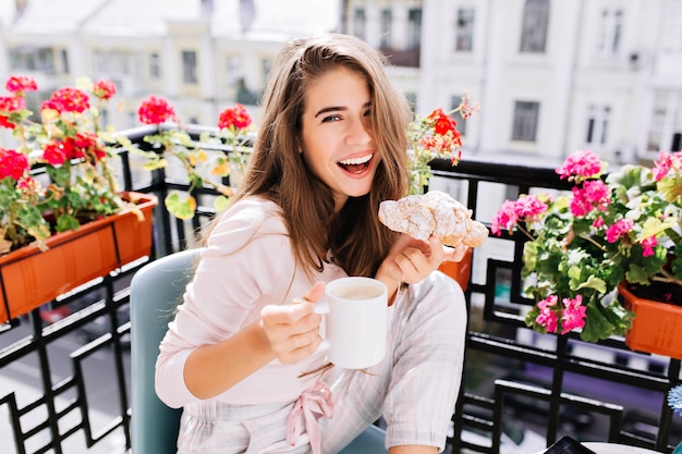 Portrait beautiful girl with long hair having breakfast on balcony in the morning in city. She holds a cup, croissant, laughing .