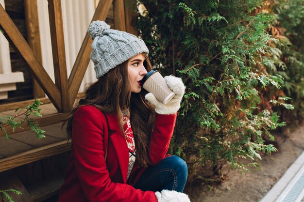 Portrait beautiful girl with long hair in red coat, knitted hat and white gloves sitting on wooden stairs near green branches outdoor. She drinks coffee and looking to side.