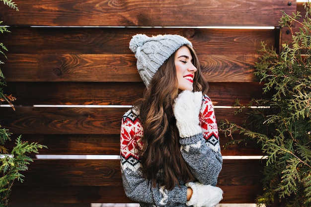 Free photo portrait beautiful girl with long hair and red lips in warm winter clothes on wooden . she is smiling to side and keeps eyes closed.