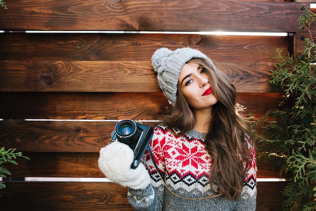 Free photo portrait beautiful girl with red lips in knitted hat and gloves holding camera on wooden .