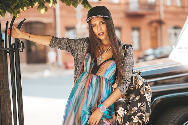 Portrait of beautiful glamor brunette teenager model in summer hipster clothes and bag. Girl posing in the street. Woman in cap