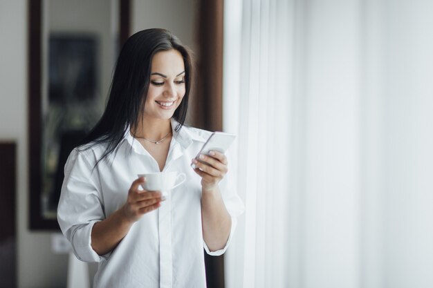 Portrait of a beautiful happy brunette girl in the morning with coffee and a phone near the window.