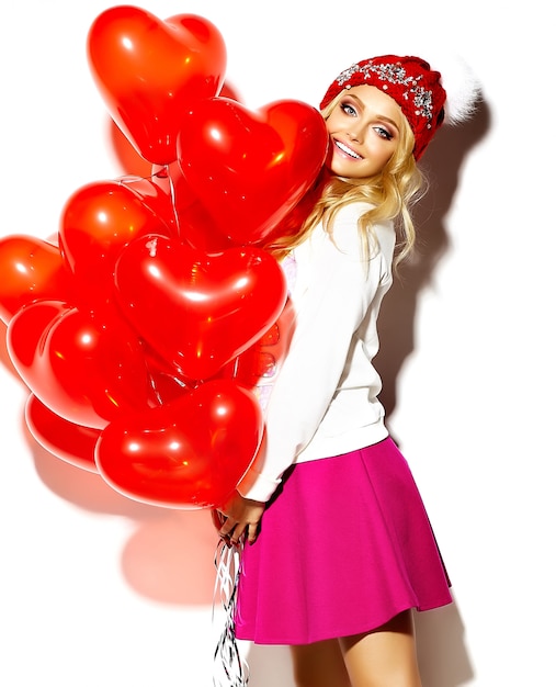 Free Photo portrait of beautiful happy sweet cute smiling blonde woman woman in casual hipster clothes, in pink skirt and winter warm beanie with red heart balloons in hands