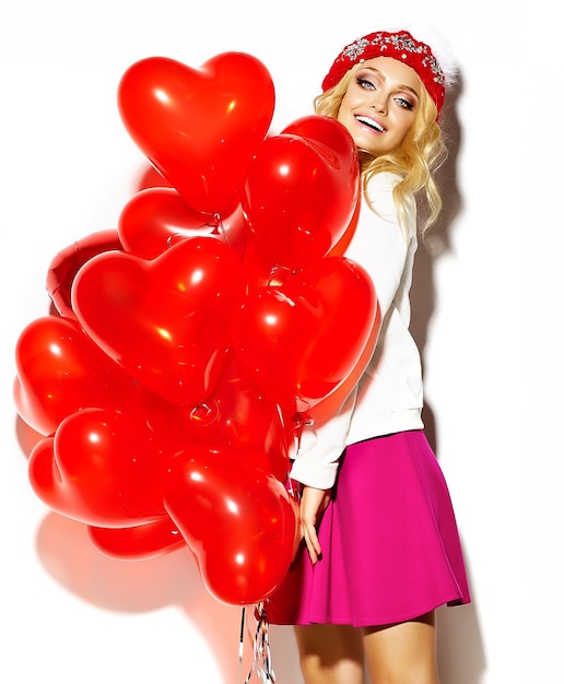 Free photo portrait of beautiful happy sweet cute smiling blonde woman woman in casual hipster clothes, in pink skirt and winter warm beanie with red heart balloons in hands