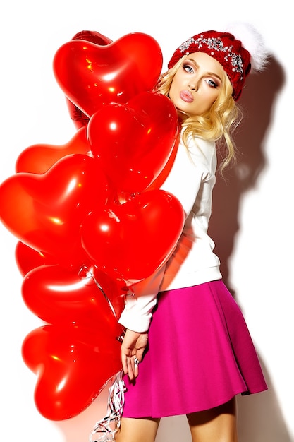 Free photo portrait of beautiful happy sweet cute smiling blonde woman woman in casual hipster clothes, in pink skirt and winter warm beanie with red heart balloons in hands