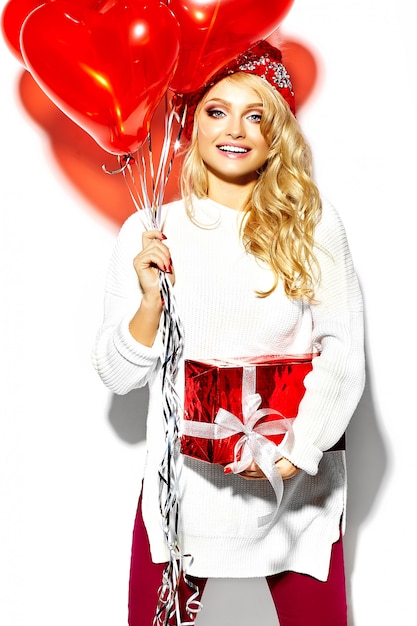 Free photo portrait of beautiful happy sweet smiling blonde woman girl holding in her hands big christmas gift box and heart balloons in casual red hipster winter clothes, in white warm sweater