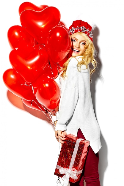 Free photo portrait of beautiful happy sweet smiling blonde woman girl holding in her hands big christmas gift box and heart balloons in casual red hipster winter clothes, in white warm sweater