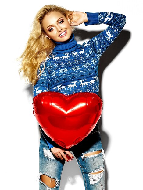 portrait of beautiful happy sweet smiling blonde woman woman holding in her hands big red heart balloon in casual hipster warm winter clothes, in blue sweater