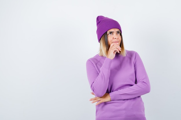 Portrait of beautiful lady keeping hand on chin in sweater, beanie and looking pensive front view