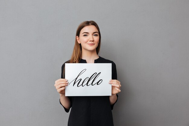 Portrait of beautiful lady standing and holding postcard while joyfully looking in camera on gray background