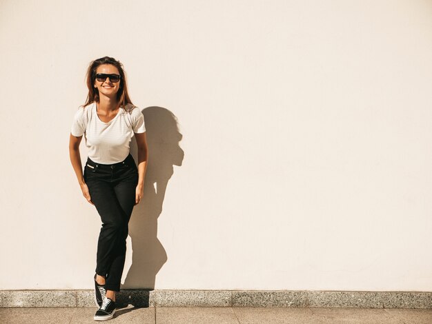 Portrait of beautiful model in sunglasses. Female dressed in summer hipster white T-shirt and jeans. Trendy woman posing near wall in the street