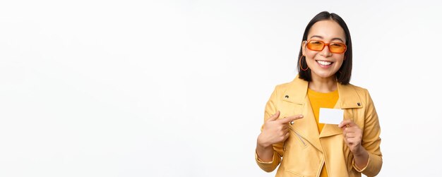 Portrait of beautiful modern asian girl in sunglasses smiling happy showing credit card standing over white background copy space