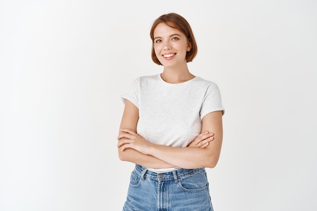 Portrait of beautiful natural female model with short hair, wearing t-shirt with jeans, cross arms on chest and smile  with happy emotion, white wall