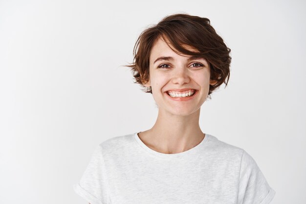 Portrait of beautiful natural girl without makeup, smiling happy , standing in t-shirt against white wall